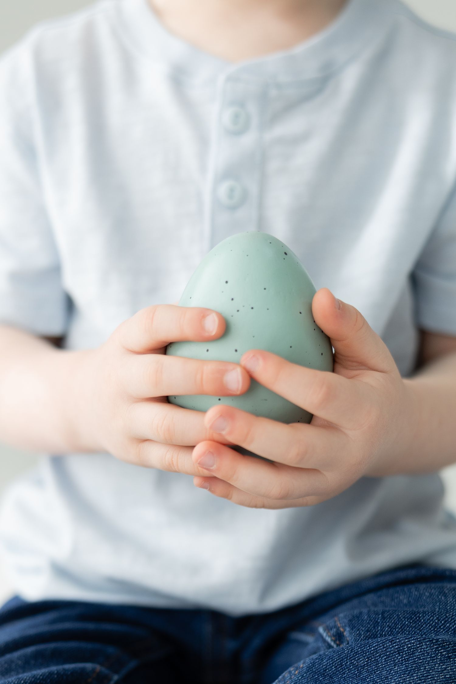 Young child in white shirt holding a large speckled mint green Easter egg with both hands.