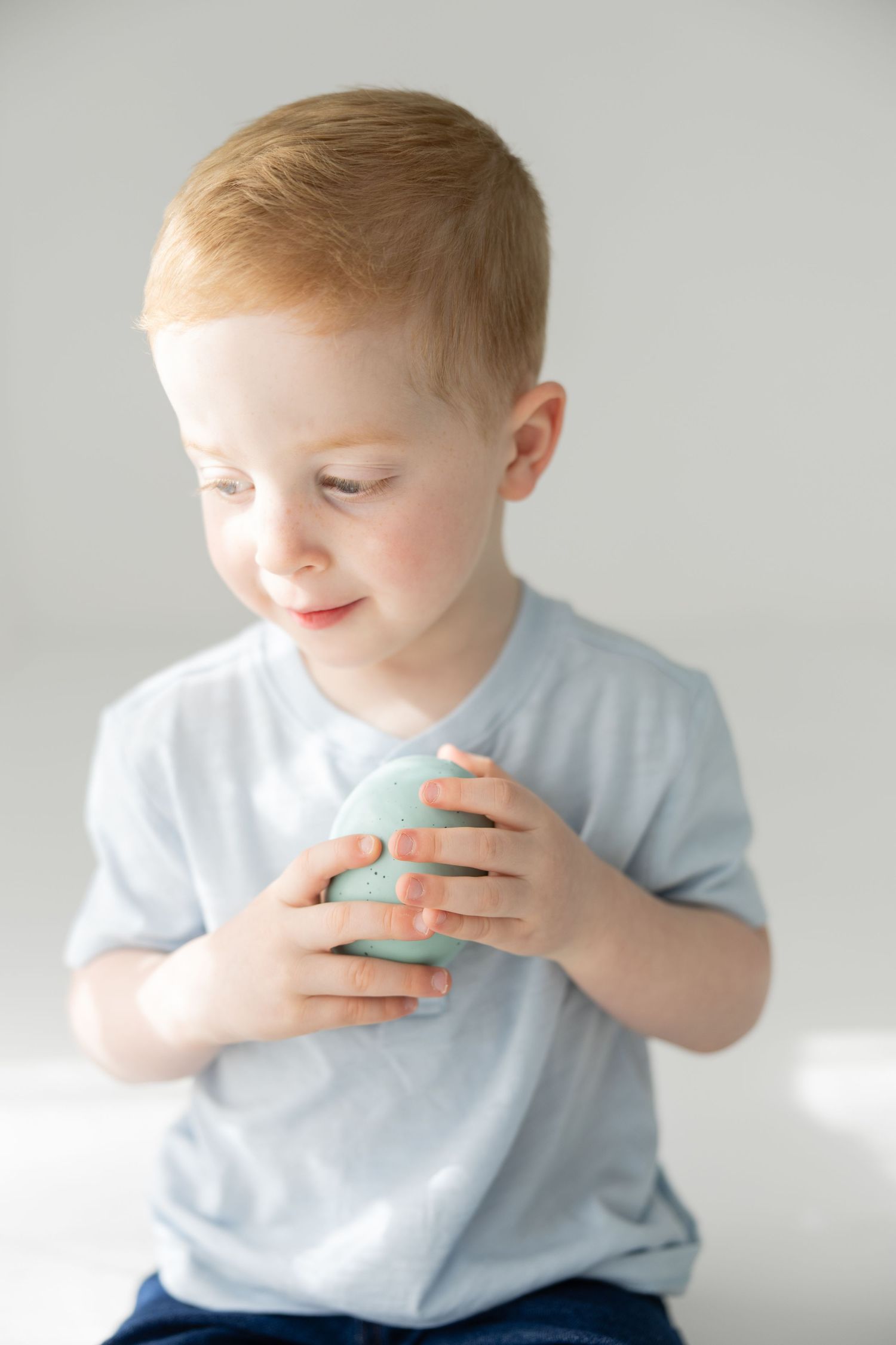 Cute redhead toddler boy holding a mint green Easter egg, wearing a light blue shirt.