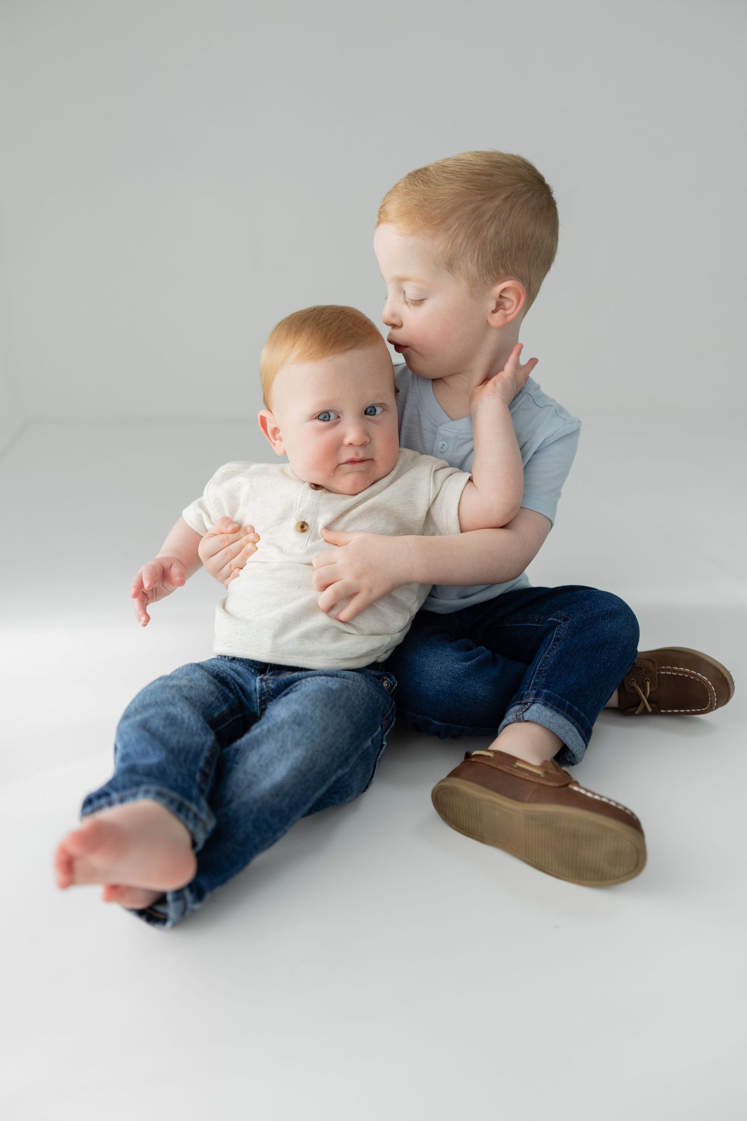 Older toddler boy kissing baby sibling on head while sitting together on white background.