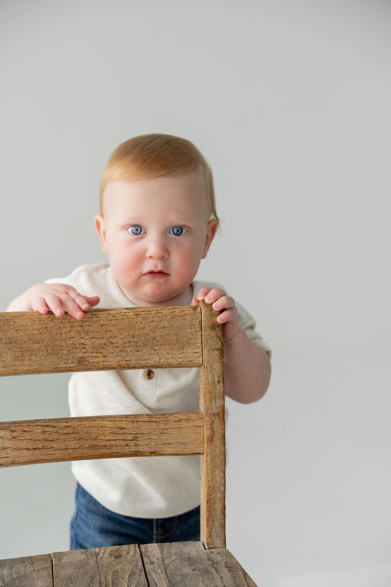Adorable blue-eyed redhead baby standing behind rustic wooden chair in white studio setting.