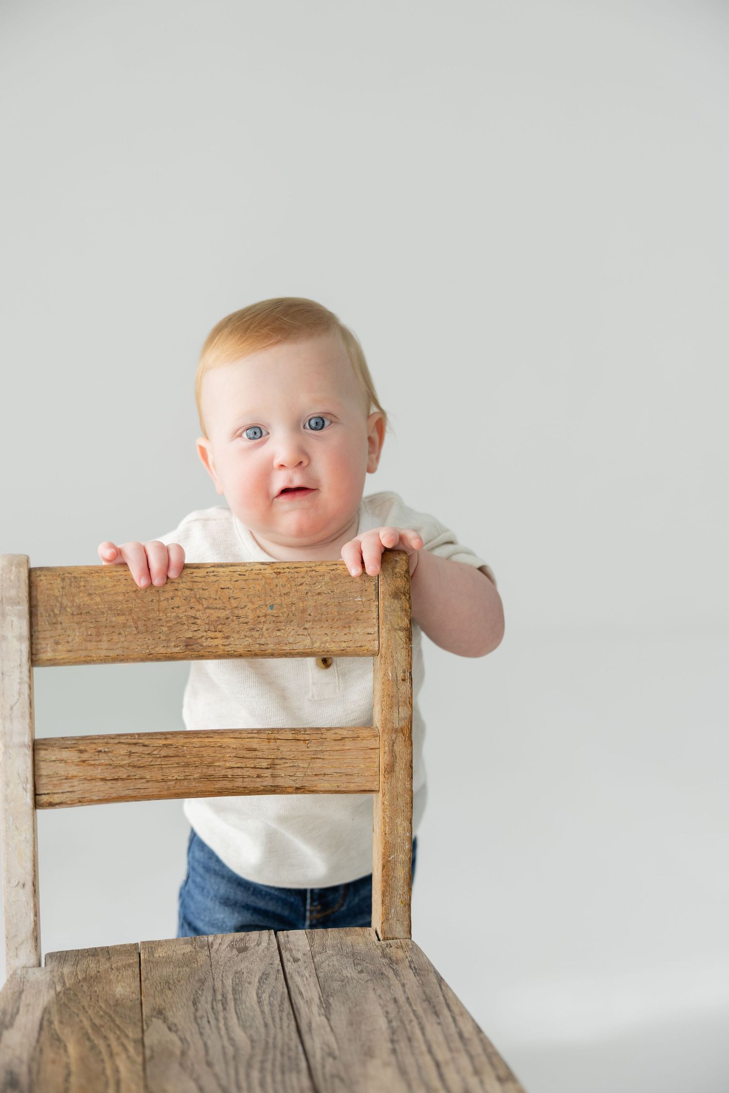 Adorable blue-eyed baby in white shirt and jeans standing behind a rustic wooden chair against a light background.