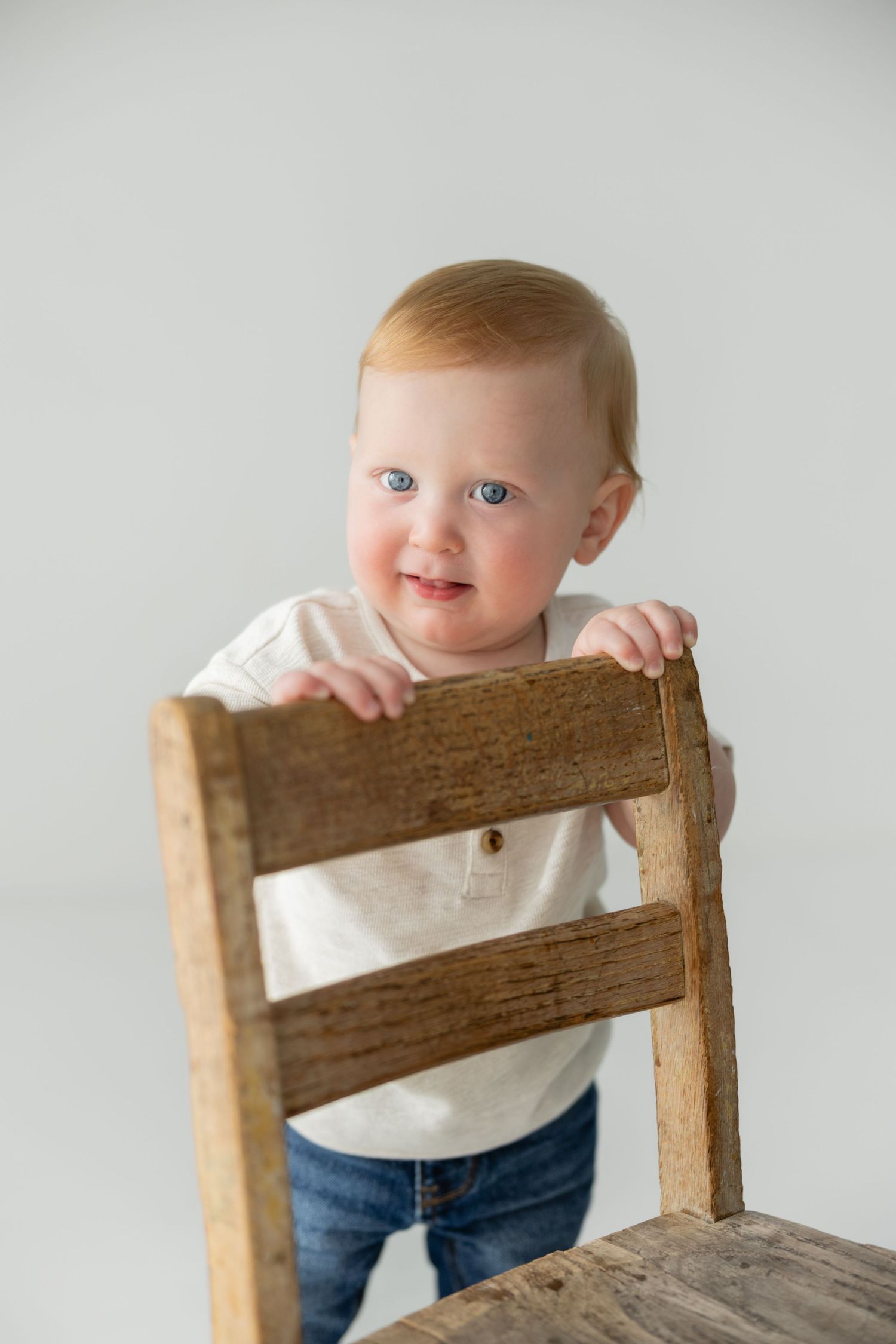 A smiling baby with blue eyes stands holding a rustic wooden chair against a white background.