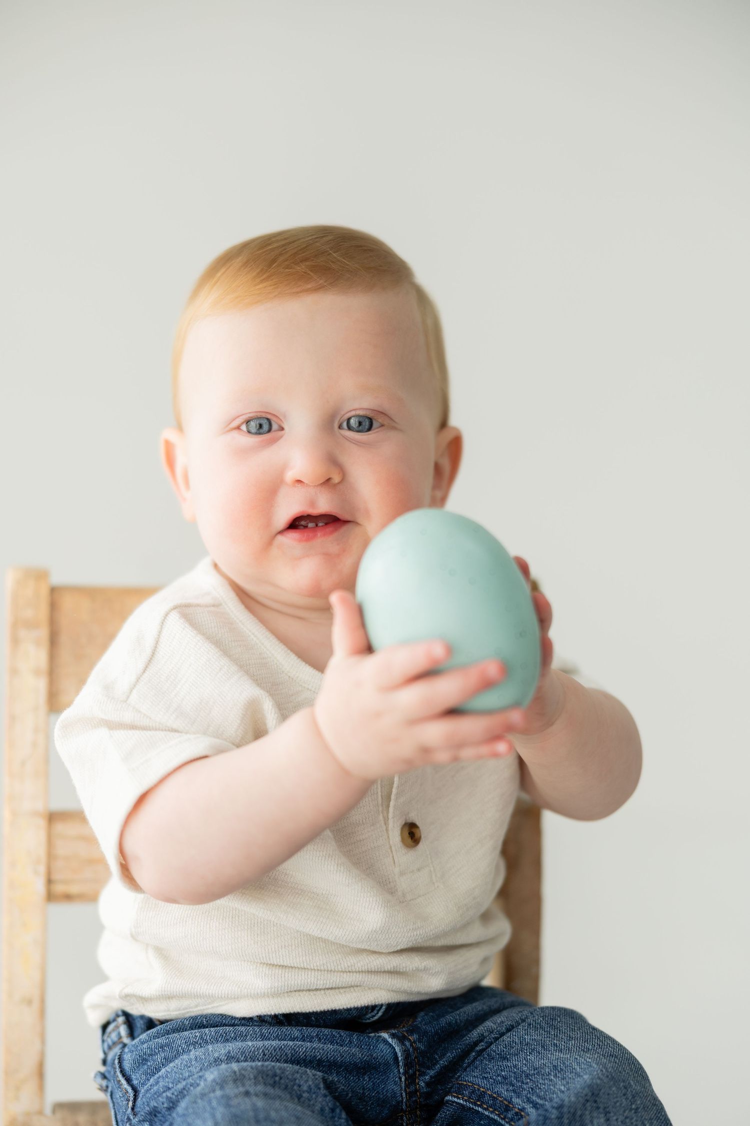 Red-haired baby with blue eyes holding a light blue Easter egg while sitting on a wooden chair.