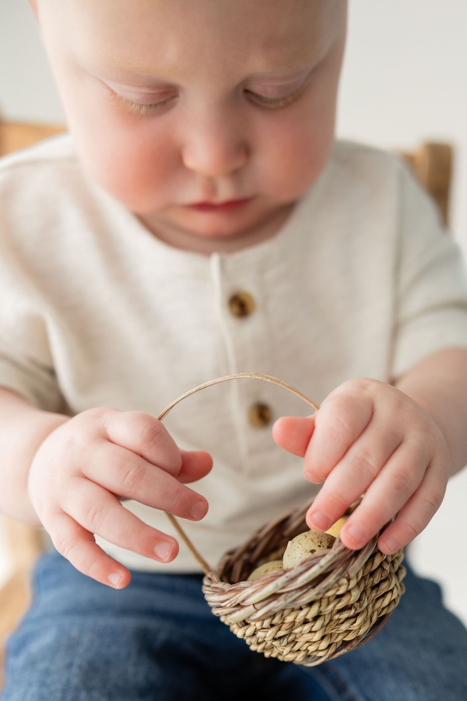 Curious toddler in linen shirt holding a small woven basket with speckled quail eggs.
