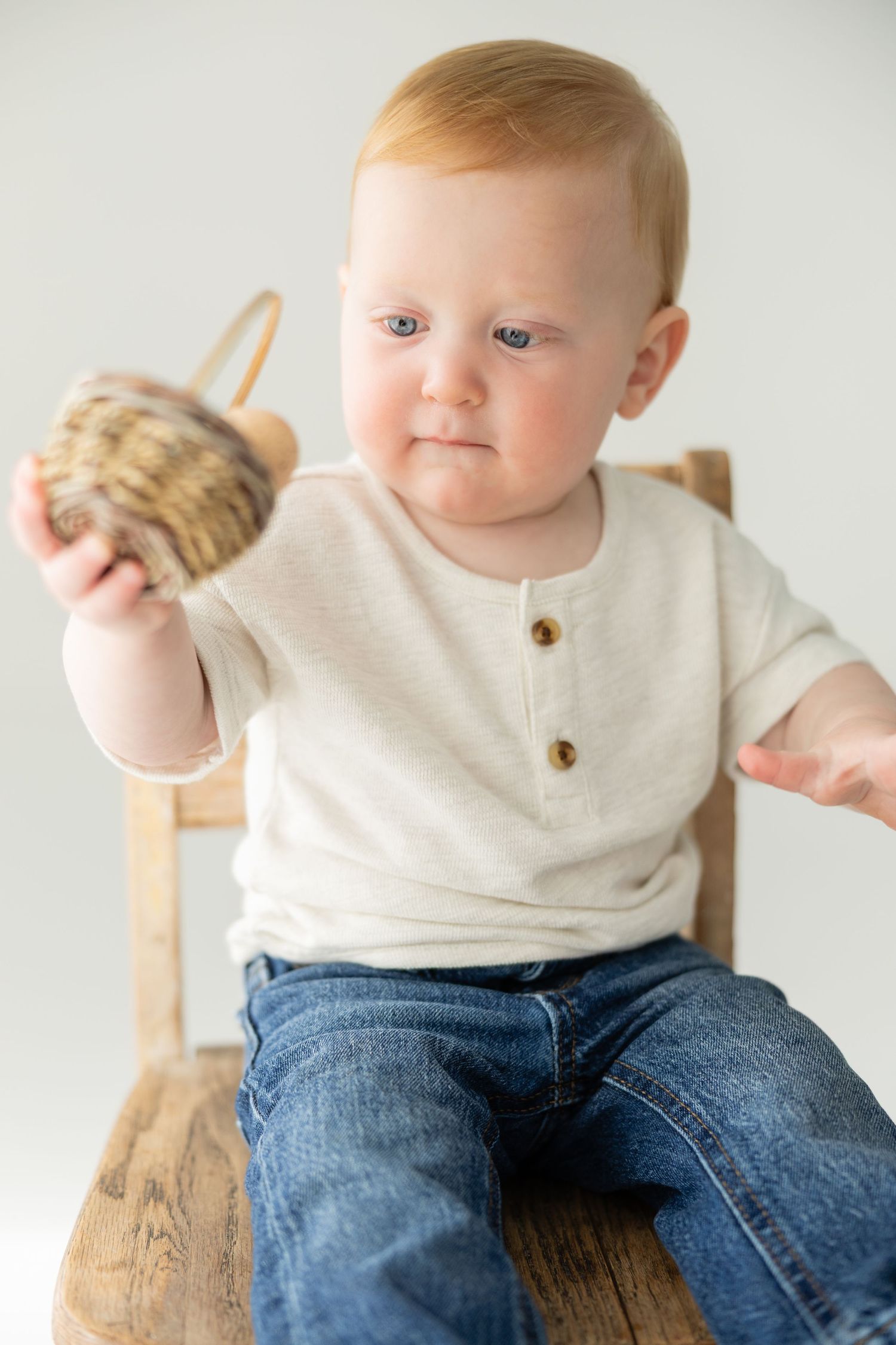 A cute baby with red hair sits on a wooden chair holding a small wicker basket, wearing a cream shirt and jeans.