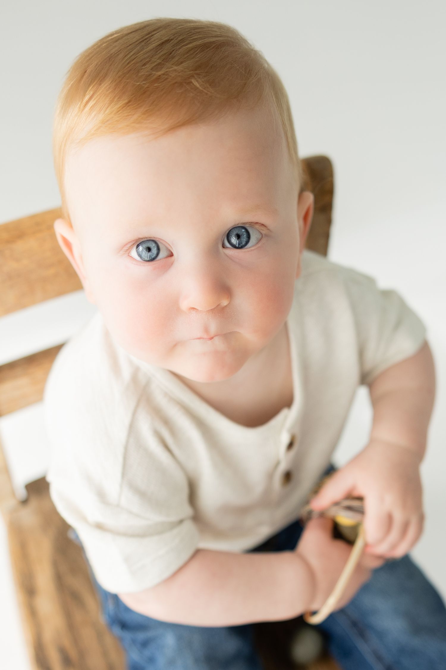 Close-up portrait of blue-eyed redhead baby sitting in wooden chair holding small toy.