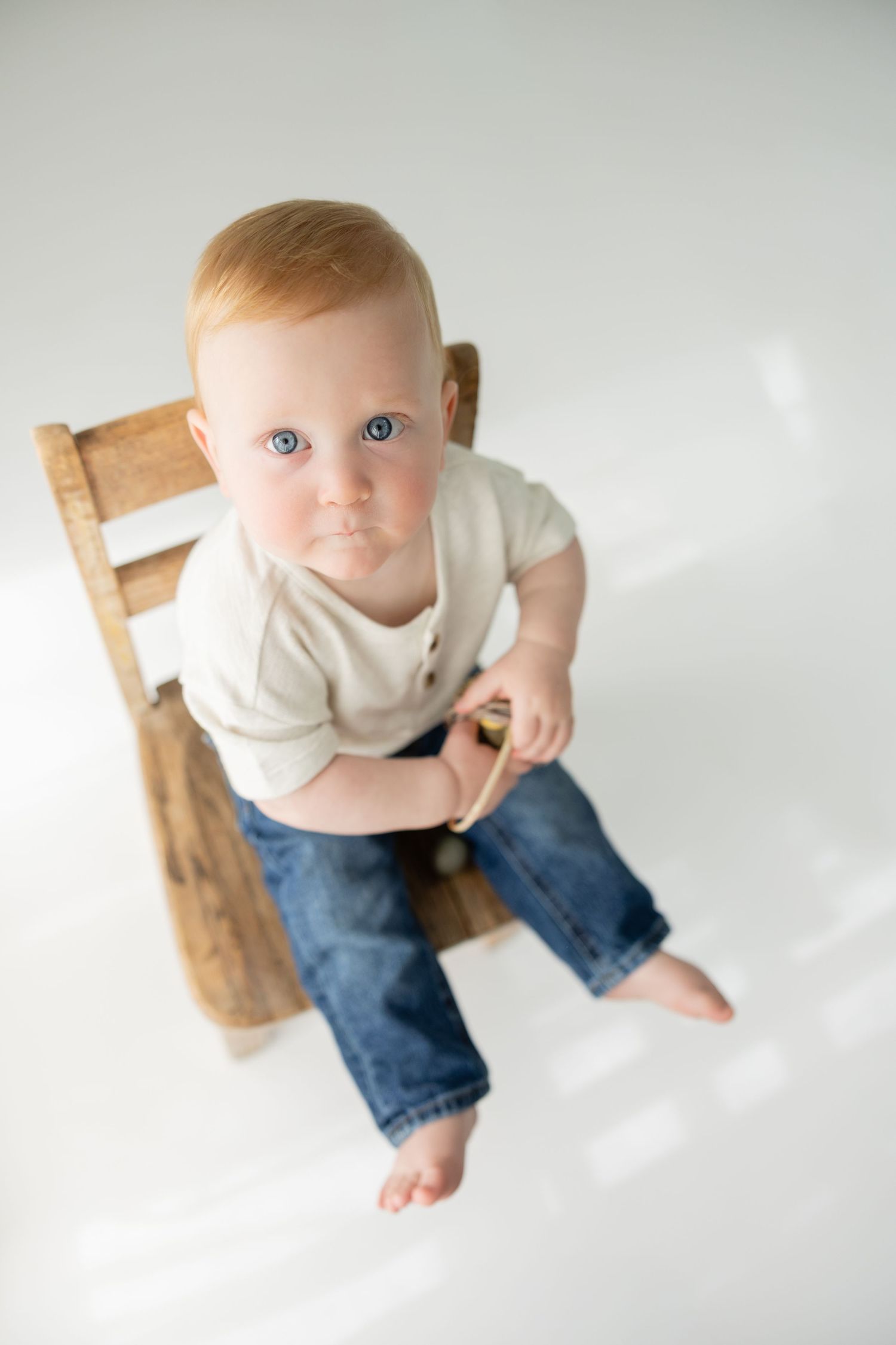 Adorable blue-eyed baby with red hair sitting on wooden chair, wearing jeans and white shirt, looking up at camera.