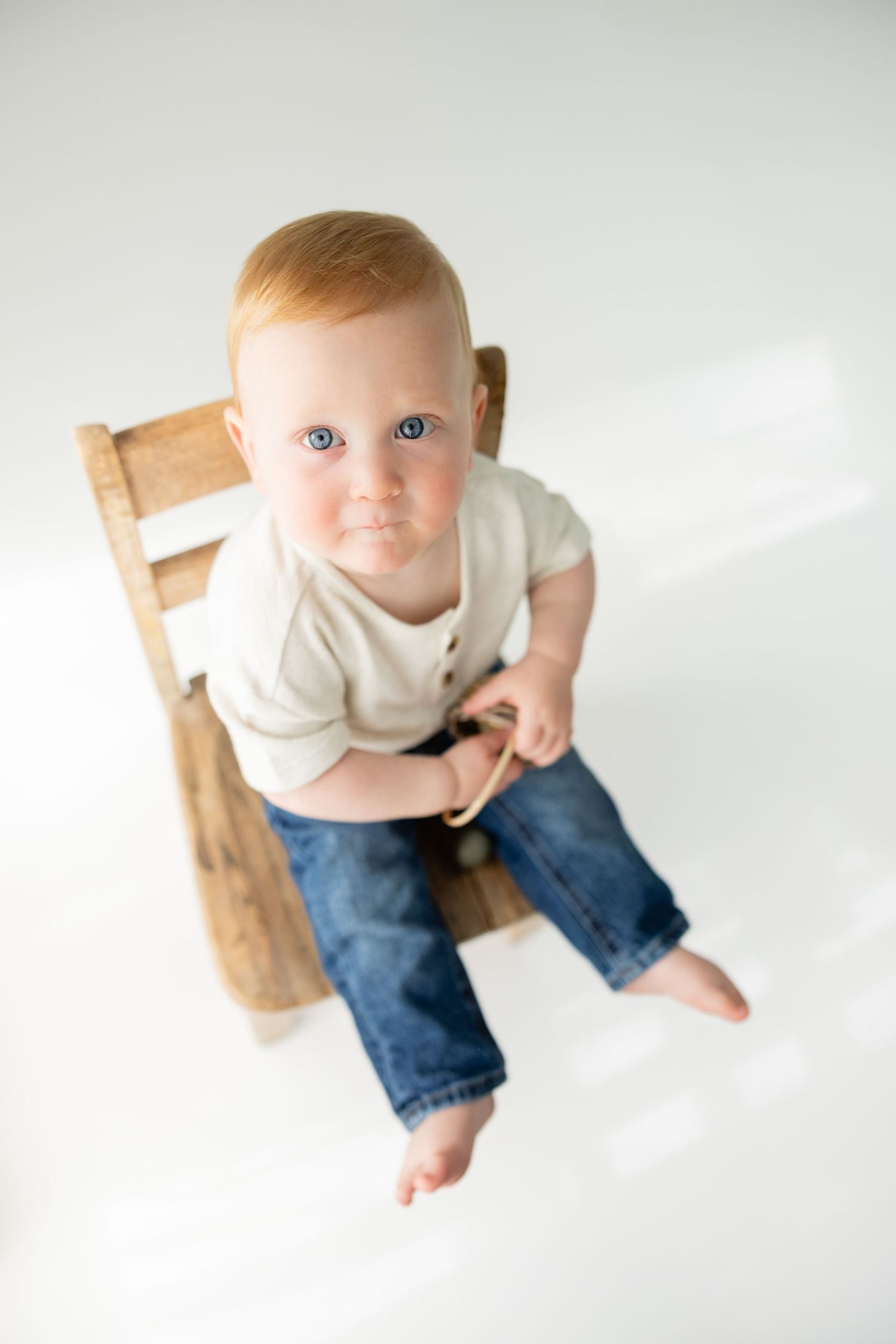 Adorable red-haired baby with blue eyes sitting on a wooden chair, wearing jeans and white onesie, looking up at camera.
