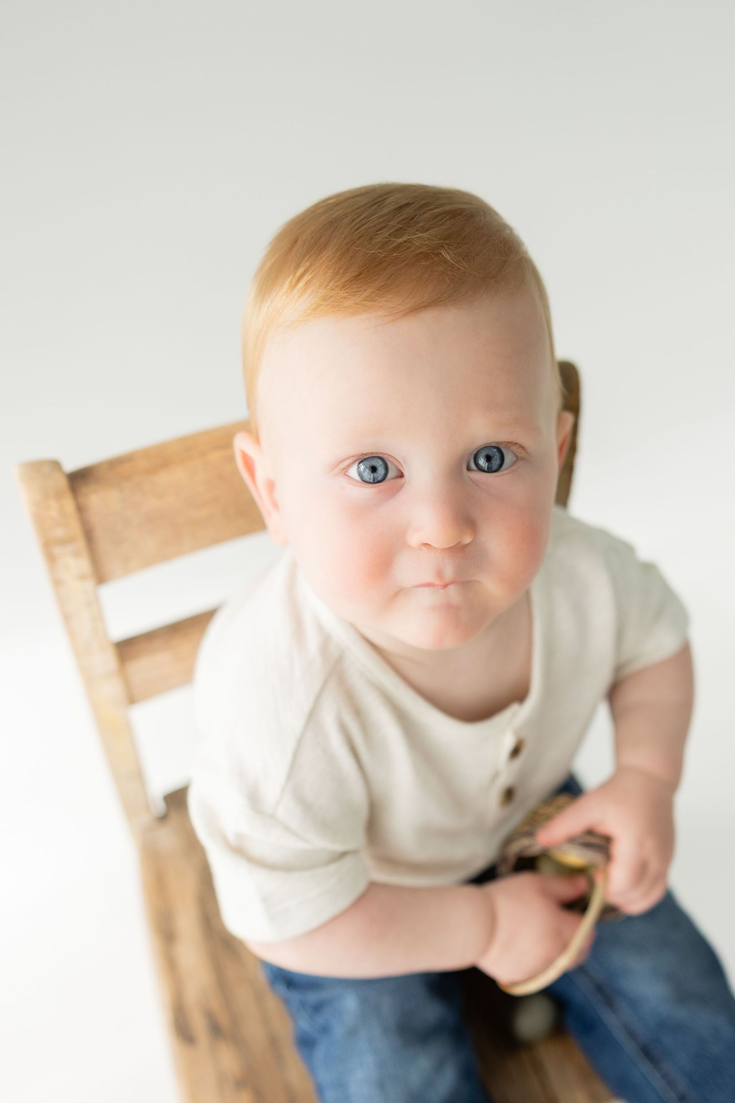Adorable red-haired baby with blue eyes sitting on a wooden chair looking up at the camera.
