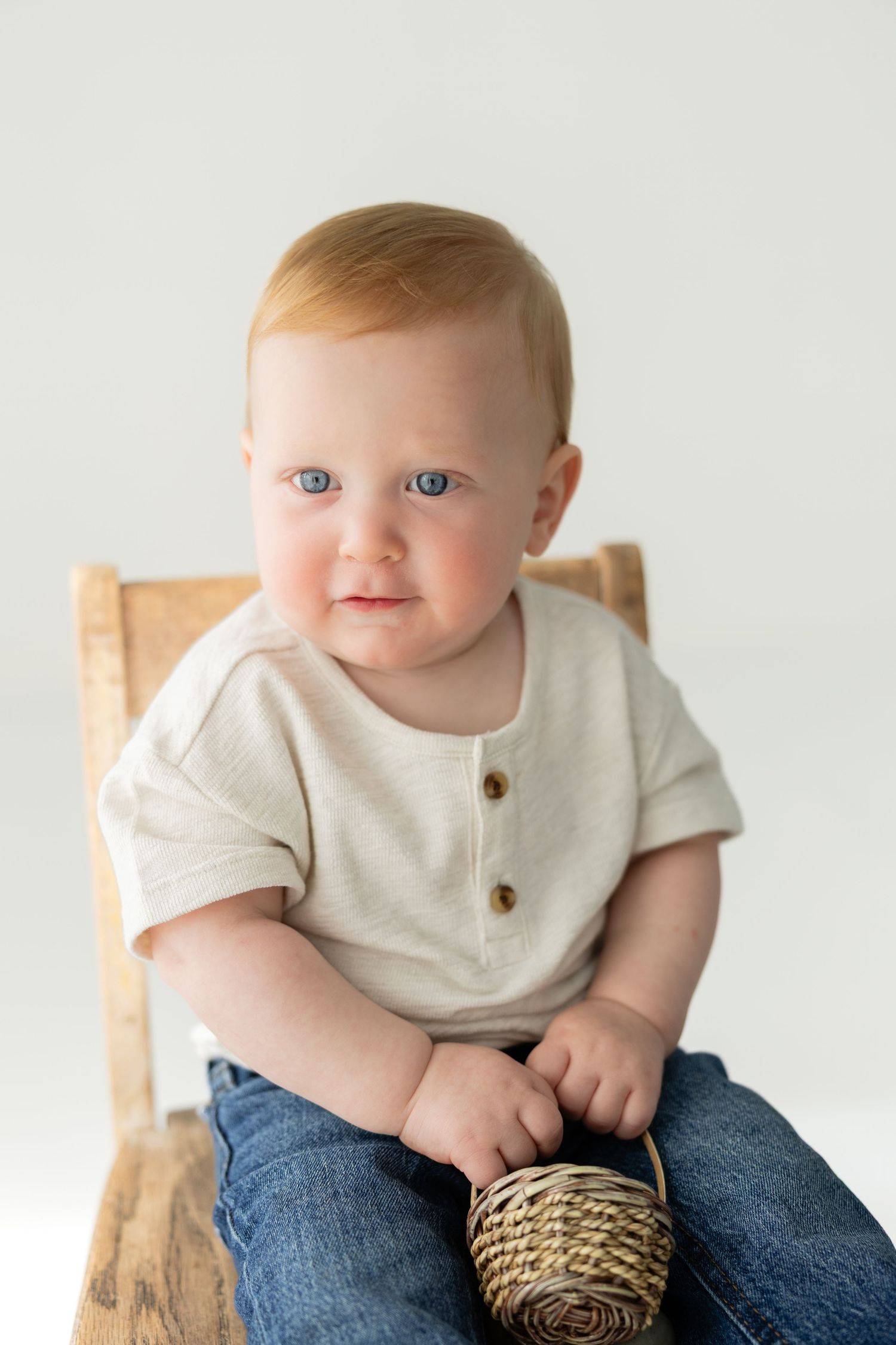 Adorable baby boy with blue eyes sits on wooden chair holding wicker basket, wearing cream henley and jeans.
