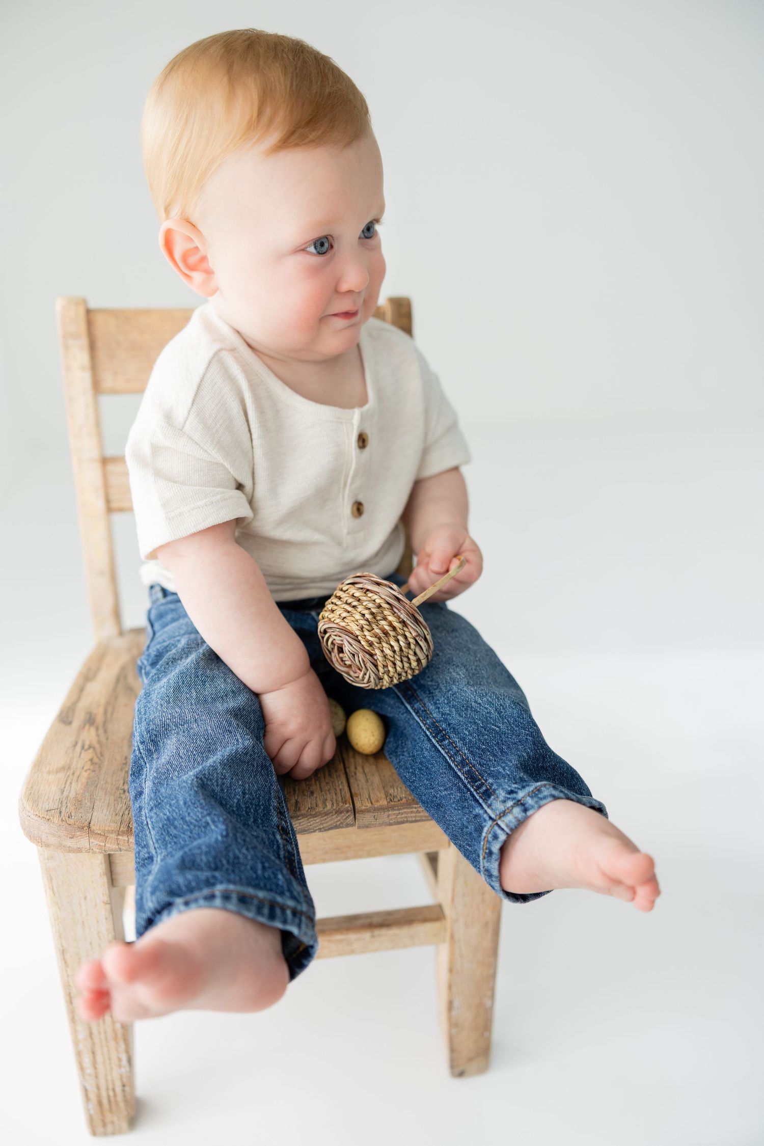 Adorable baby in jeans and cream shirt sitting on rustic wooden chair holding woven toy on white background.
