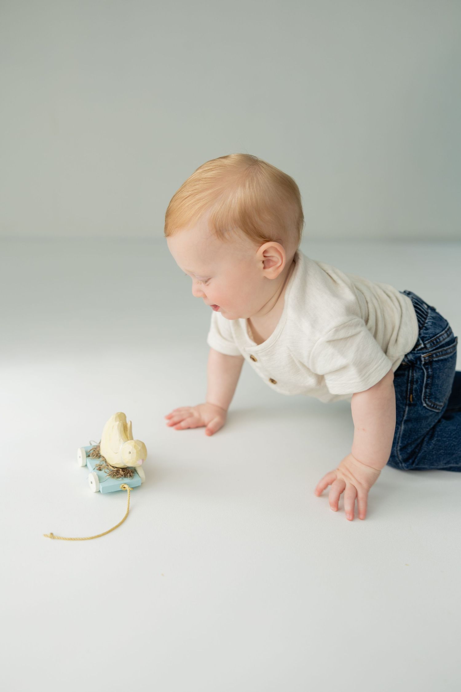 Red-haired baby in white onesie and jeans crawling toward a small toy on white floor.