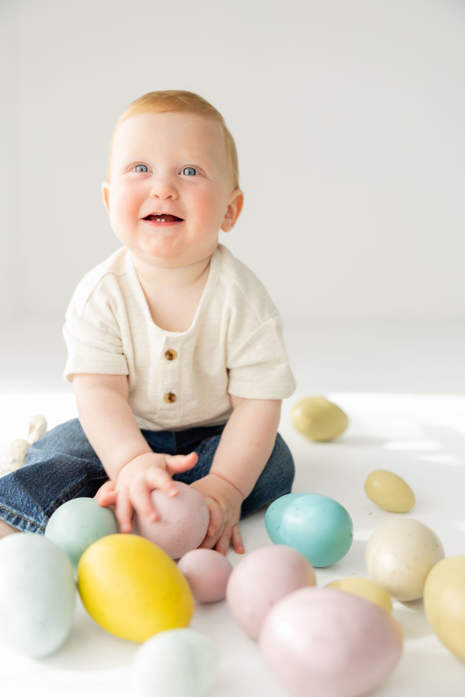 Happy red-haired baby laughing while sitting surrounded by colorful pastel Easter eggs.