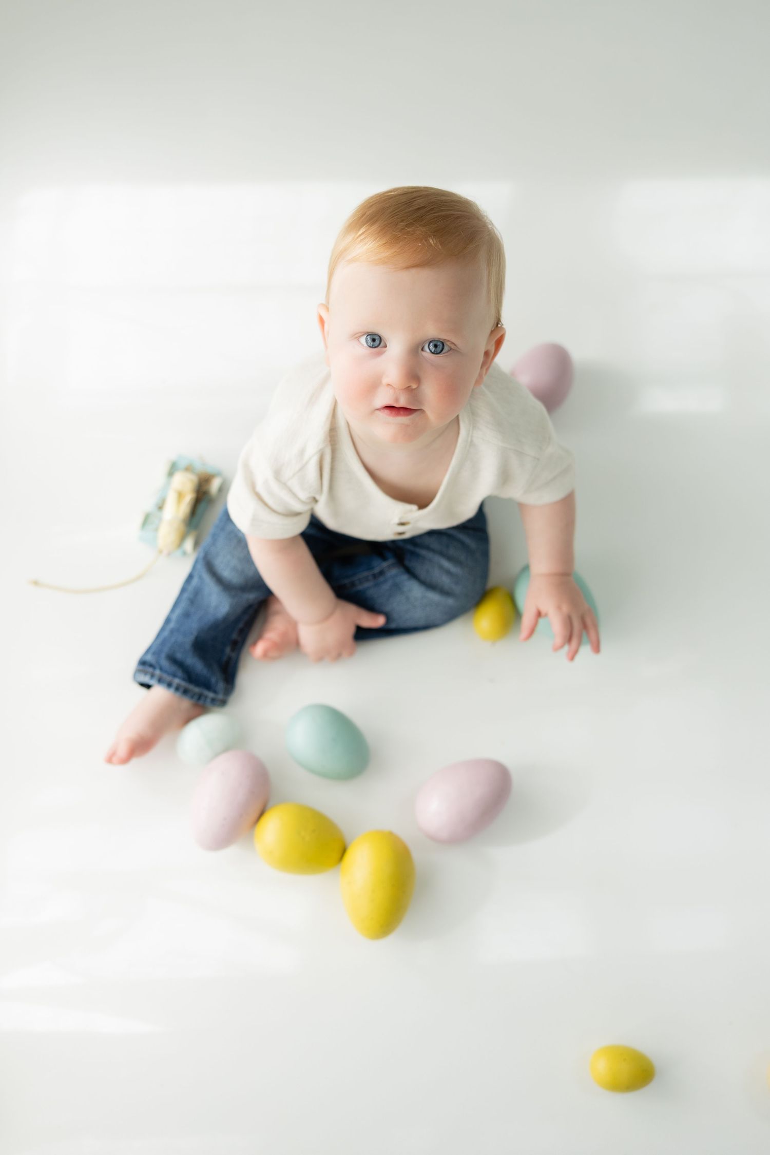 Blue-eyed red-haired baby sitting on white floor surrounded by colorful pastel Easter eggs.