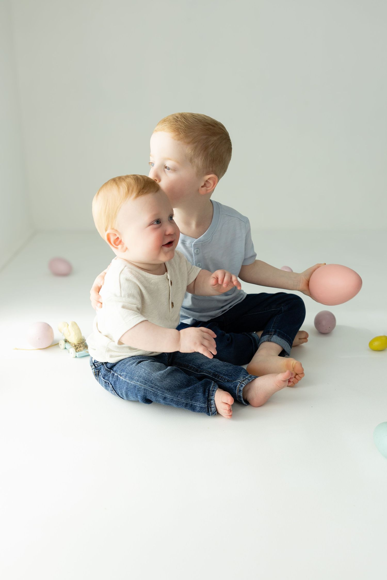 Two redhead brothers sitting together on white floor surrounded by pastel Easter eggs.