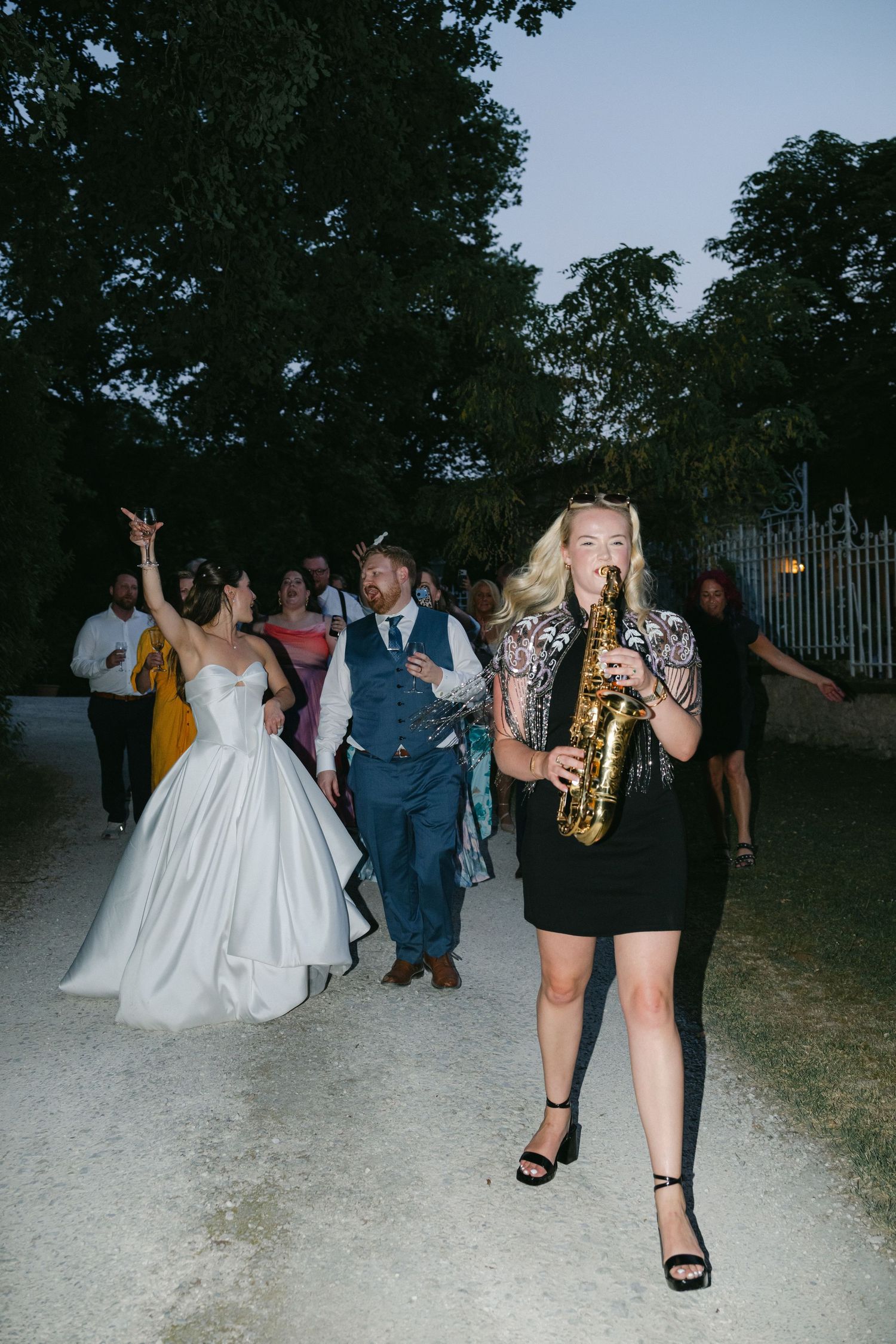 Saxophonist leads wedding procession outdoors at dusk, with bride in white gown and guests following along a garden path.