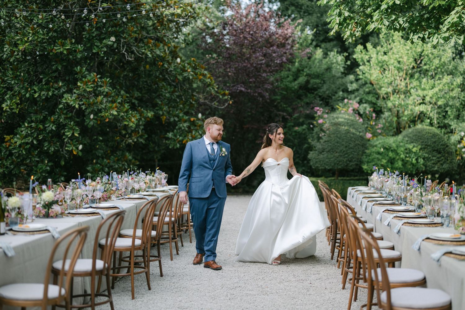 Bride and groom walk hand-in-hand between elegantly set outdoor reception tables in a lush garden wedding venue.