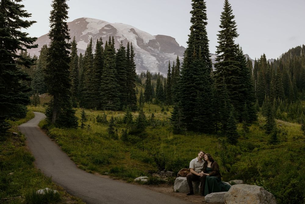 Couple sitting on rocks at Mount Rainier with evergreen trees and snow-capped mountain in background.