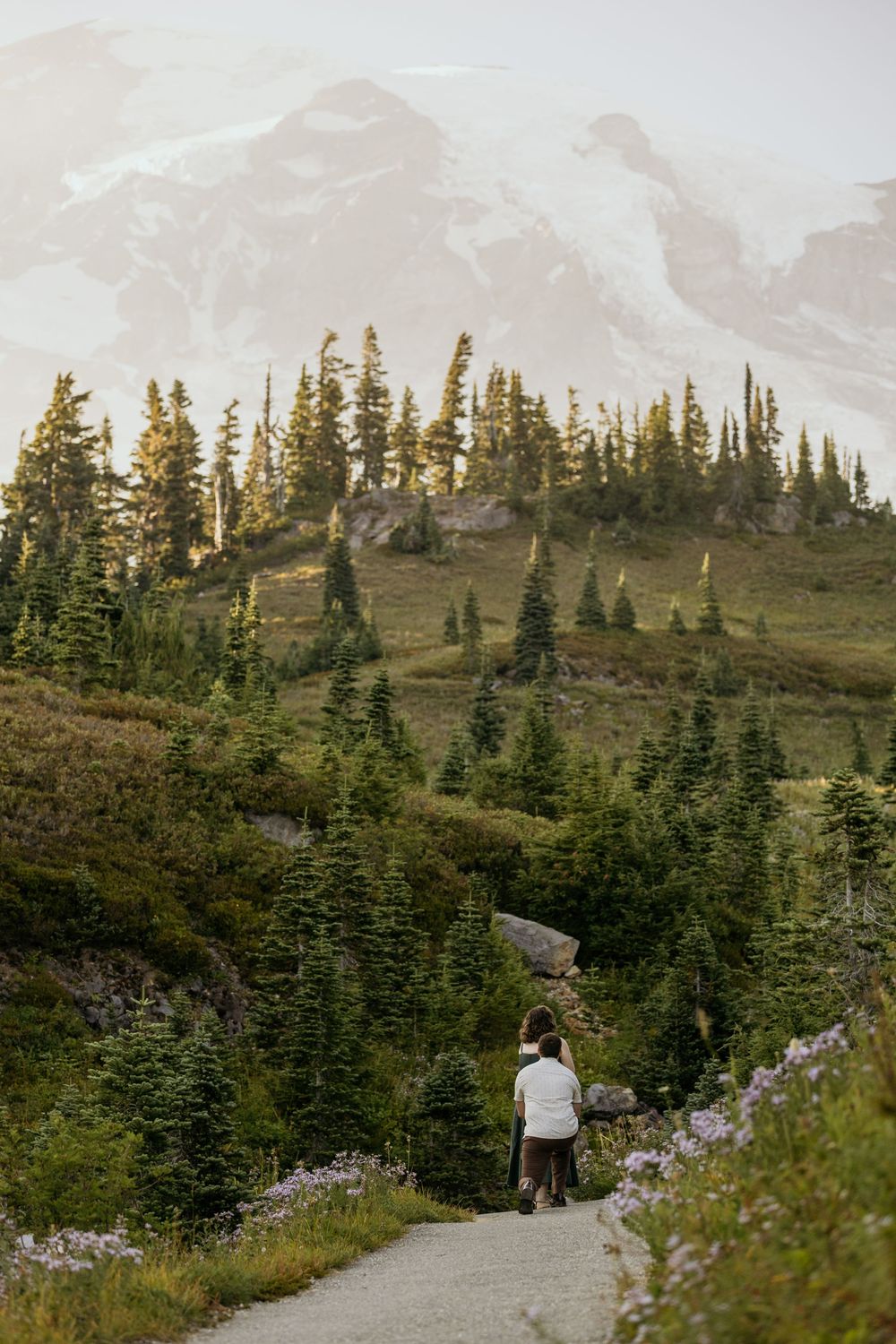 Hiker walks along a scenic mountain trail surrounded by evergreen trees with snow-capped peaks in the background.