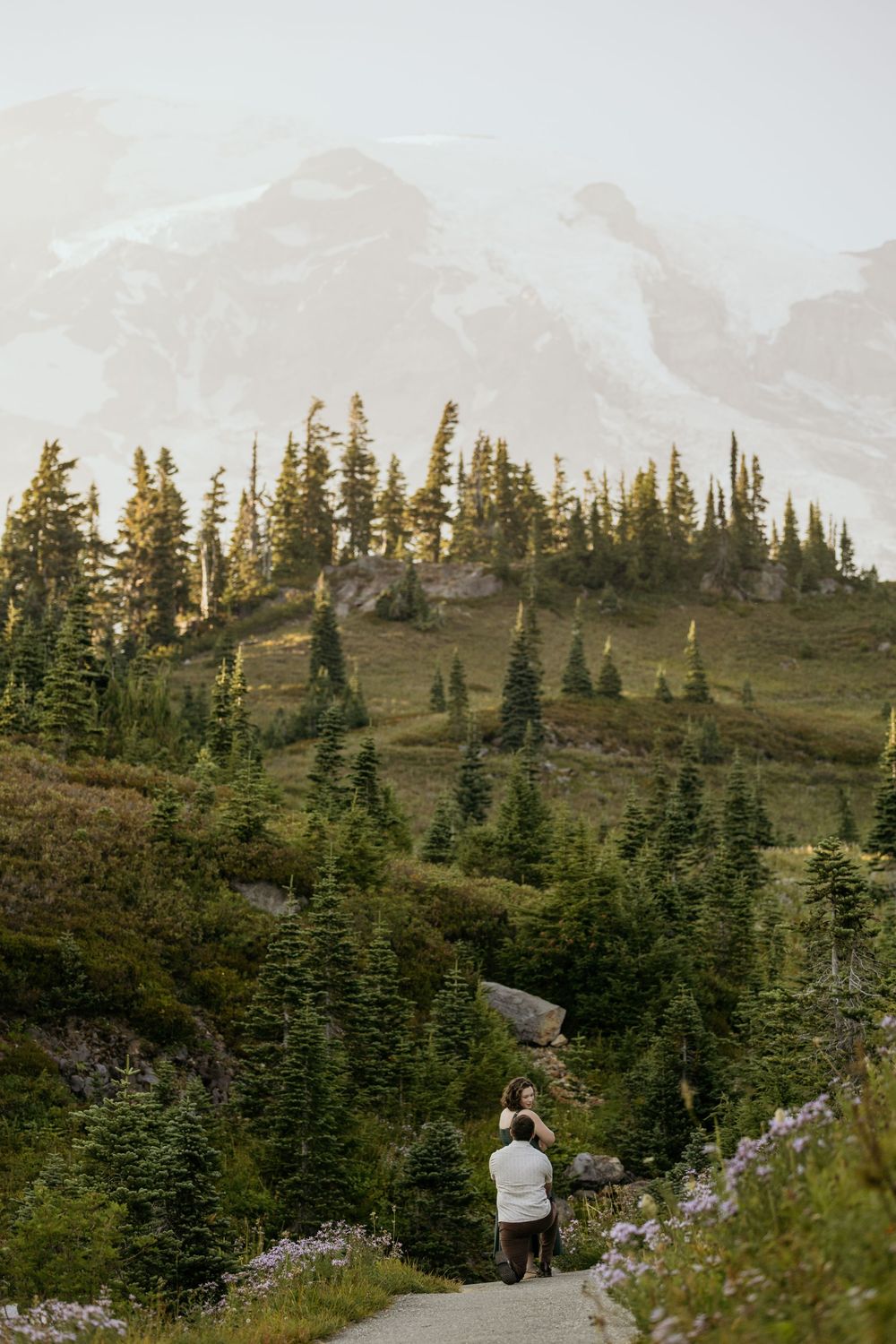 Couple walking on a mountain trail surrounded by evergreen trees with a misty peak in the background.