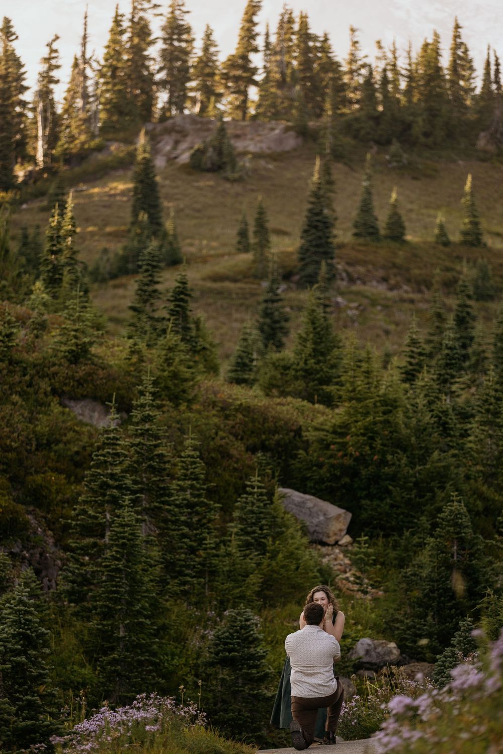Person proposing on one knee on a mountain trail surrounded by evergreen trees at golden hour.