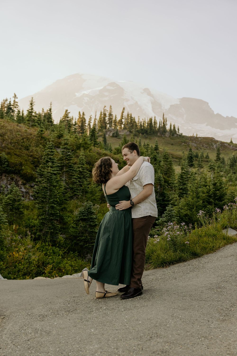 Couple embracing on a mountain trail with evergreen trees and snow-capped peak backdrop.