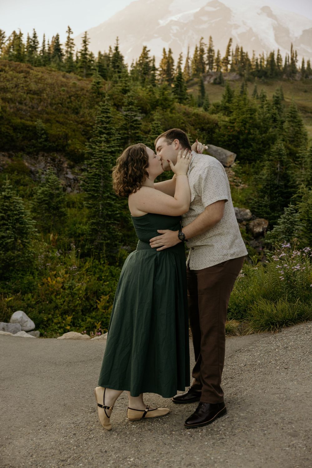 Couple sharing a romantic kiss in front of evergreen trees and a snow-capped mountain, woman in green dress.