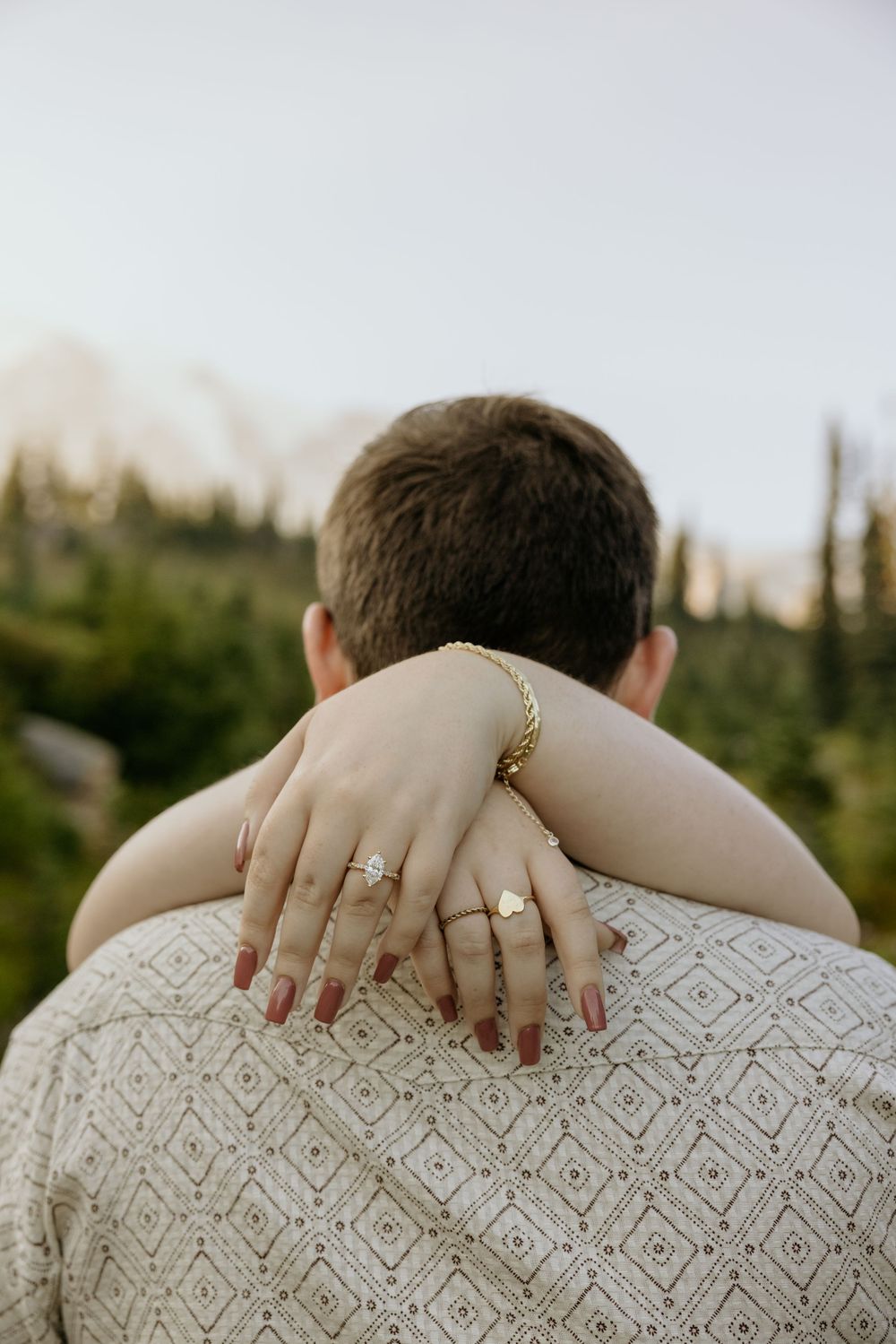 Close-up of a woman's hands with gold rings and bracelet wrapped around her partner's shoulders outdoors.
