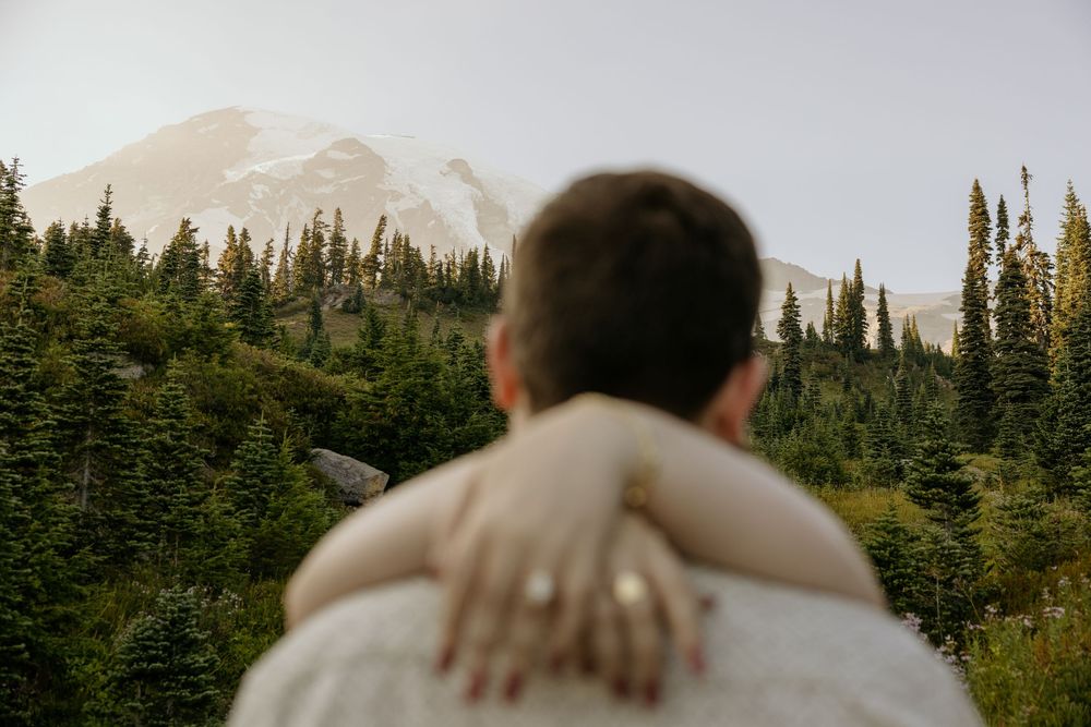 Couple embracing with mountain and evergreen forest backdrop, engagement ring visible on hand.