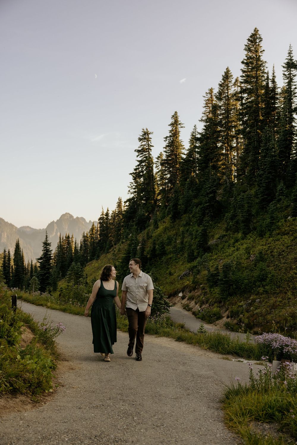 Couple walks hand-in-hand on a mountain trail surrounded by tall evergreen trees at golden hour.