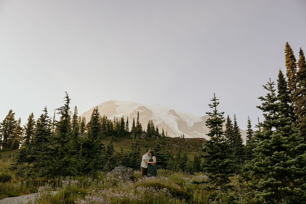 Couple embracing in a wildflower meadow with evergreen trees and Mount Rainier in the misty background.