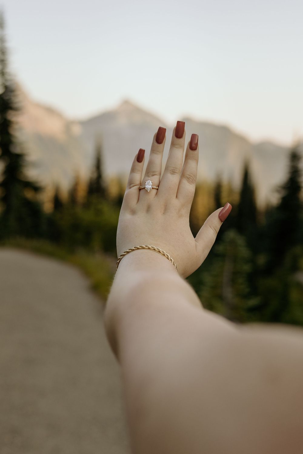 Woman showing off emerald cut diamond engagement ring with dark red nails and gold bracelet against mountain backdrop.