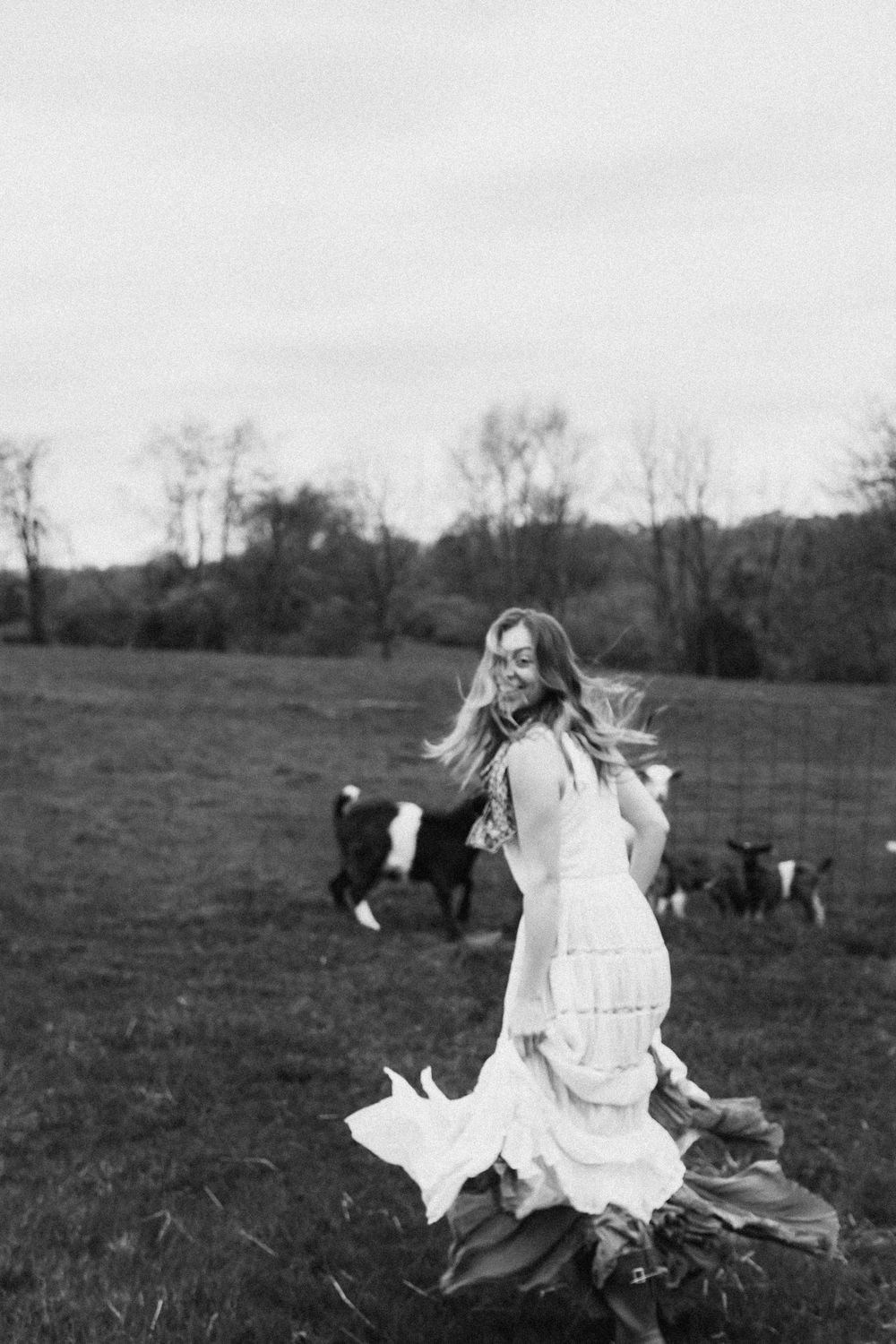 Black and white photo of a smiling woman in a white dress twirling in a field with cows grazing in the background.