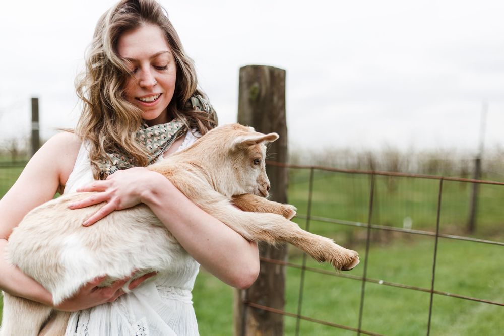 Woman in white dress holding a baby goat near a wooden fence post on a green farm.