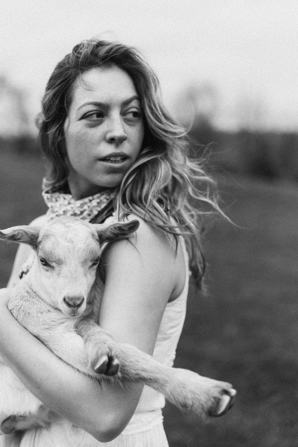 Young woman with wavy hair holds a baby goat in a black and white farm portrait photo.