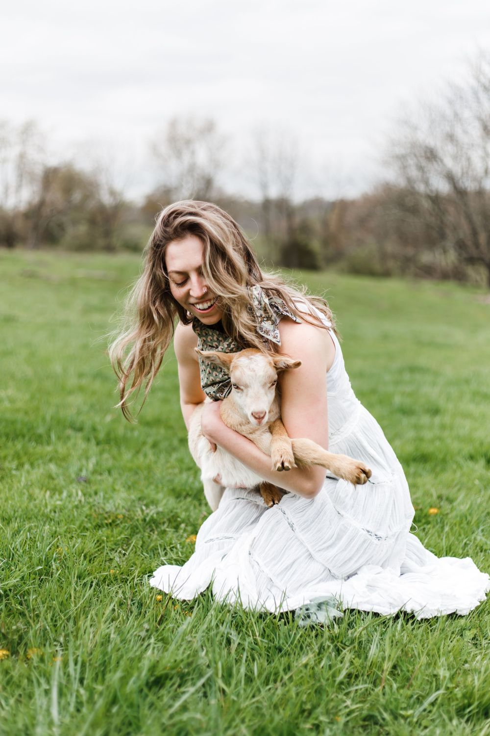 A woman in a white dress crouches on green grass, smiling while gently hugging a small baby goat.