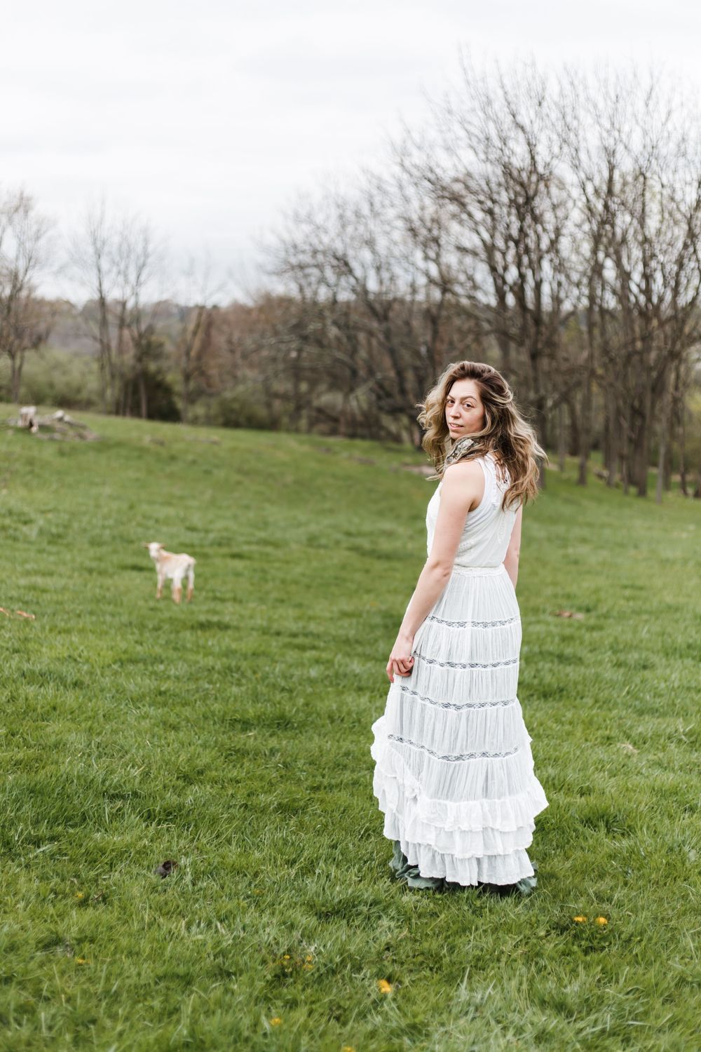 A woman in a white tiered maxi skirt stands on a green grassy field with a dog and bare trees in the background.