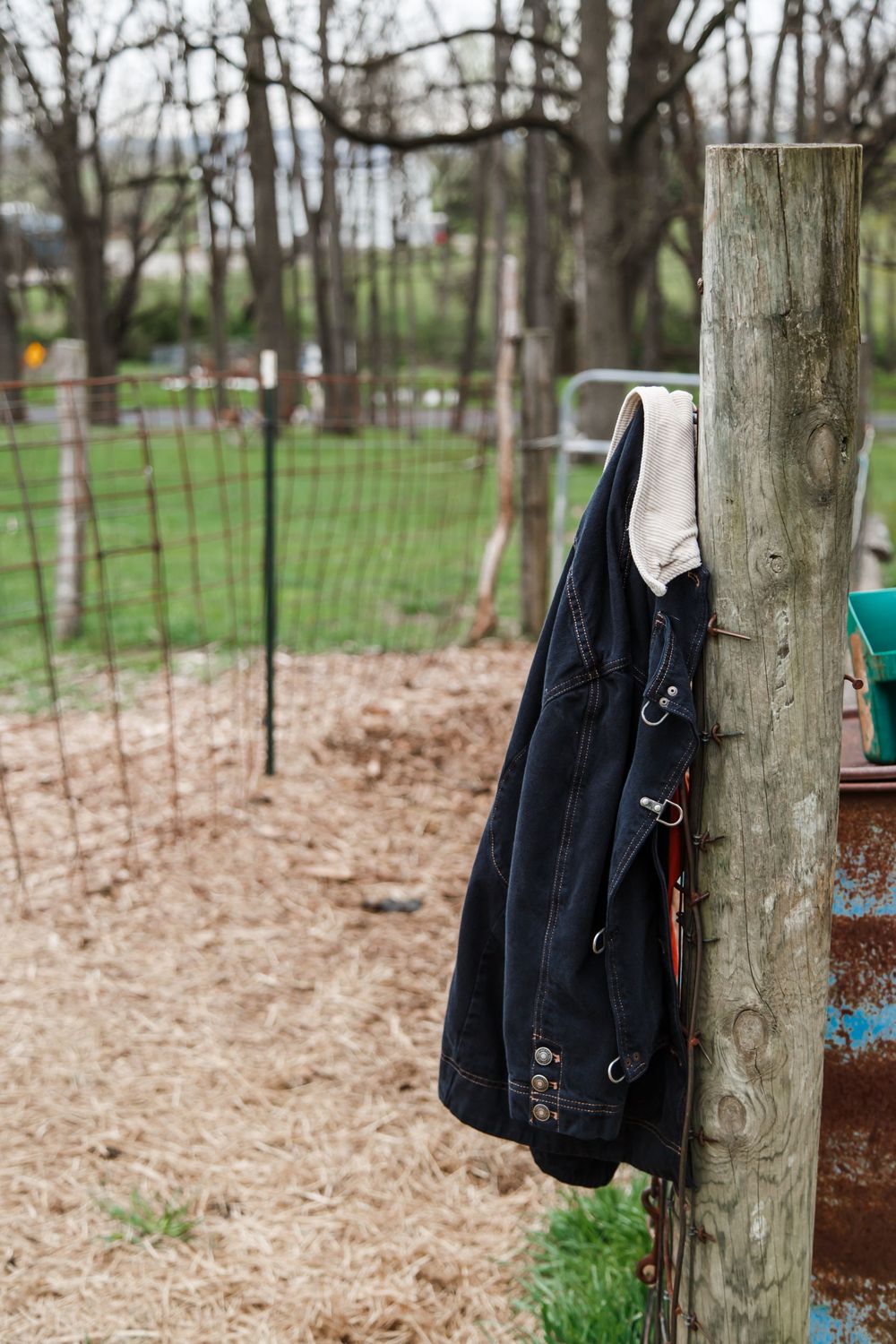 Denim jacket hanging on a wooden fence post in a rural farm setting with bare trees and wire fencing.