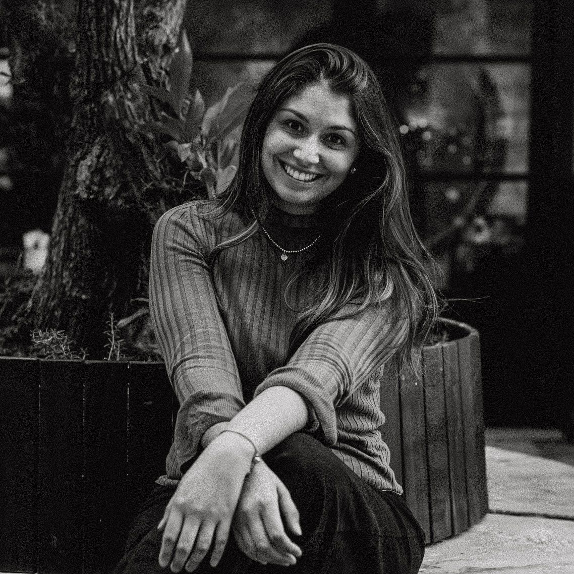 Young woman smiling and sitting outdoors in black and white portrait photography near a tree.