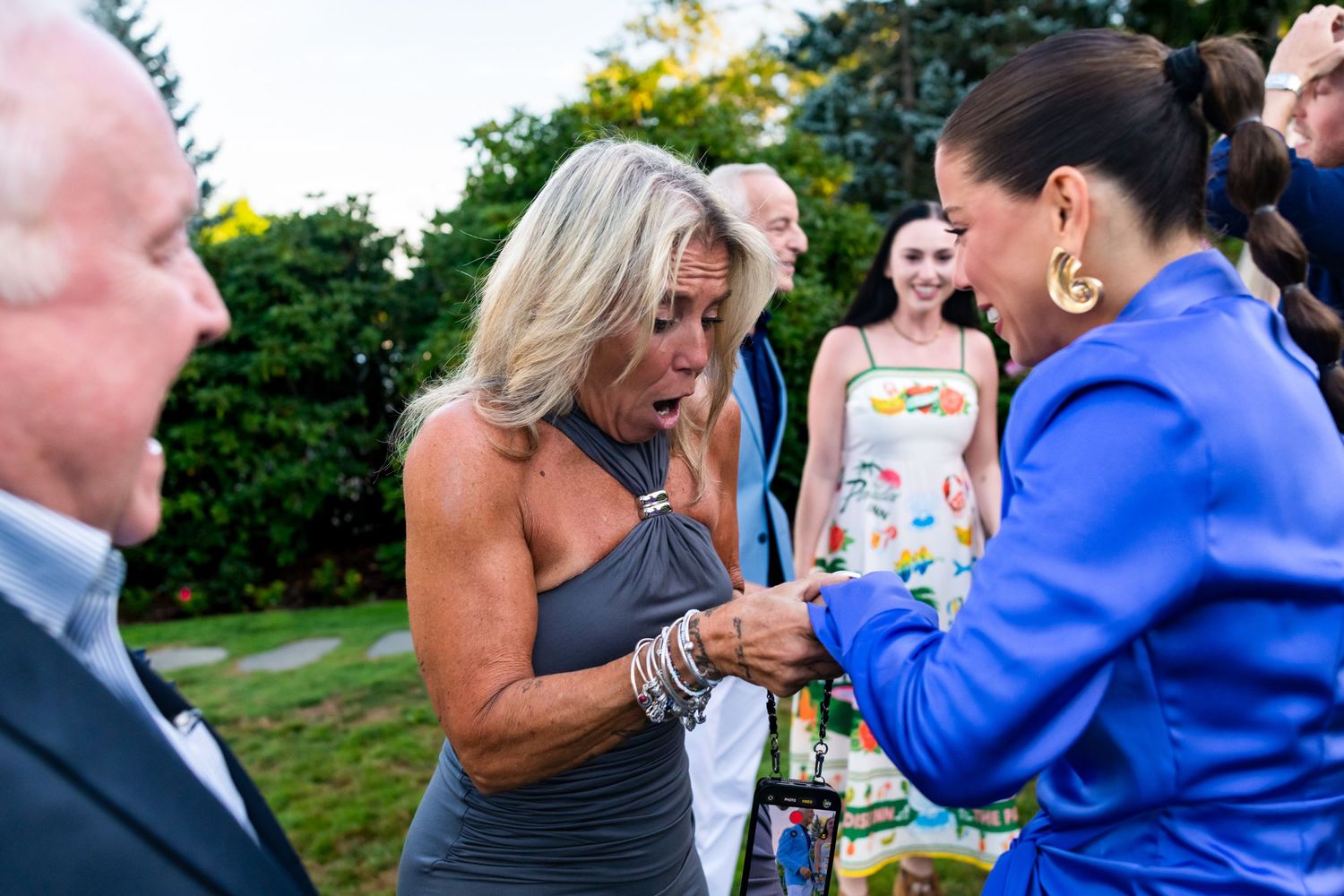 A blonde woman reacts with surprise as a woman in blue shows her something at an outdoor event.
