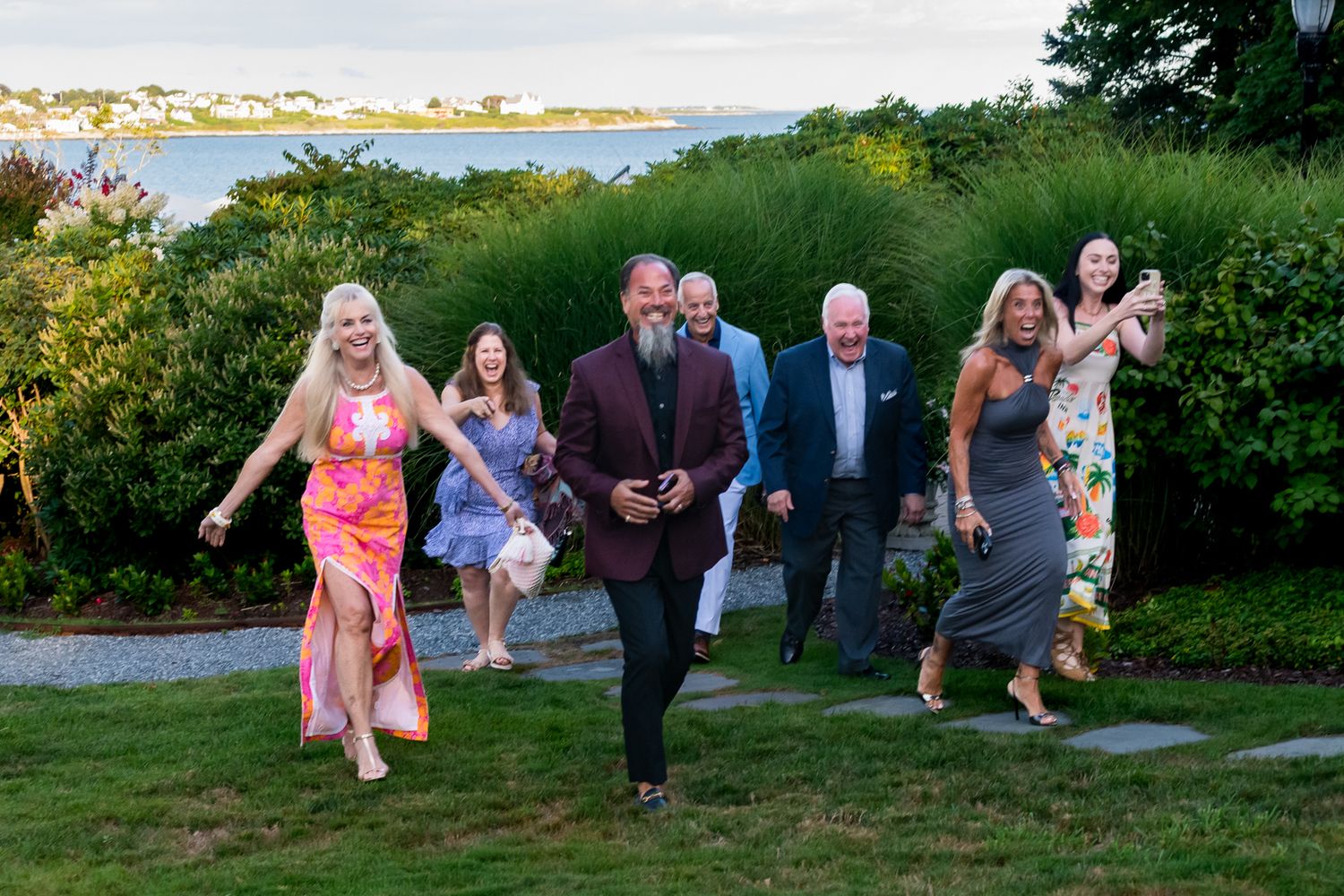 Group of elegantly dressed guests walking through garden at outdoor evening event.