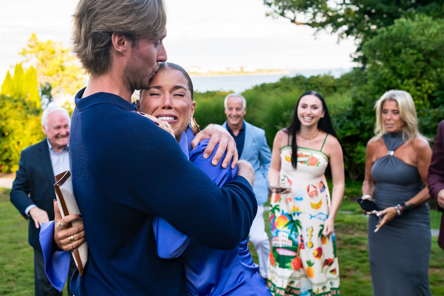 Couple sharing joyful hug surrounded by smiling guests at outdoor celebration.