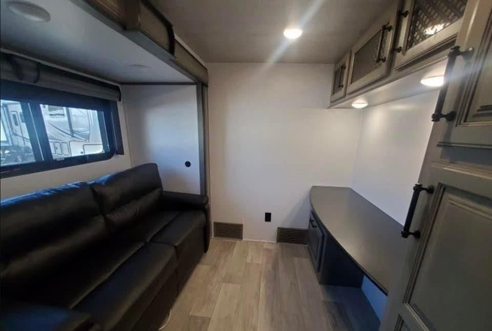 Compact RV living space with black leather sofa, fold-down desk, and overhead cabinets.