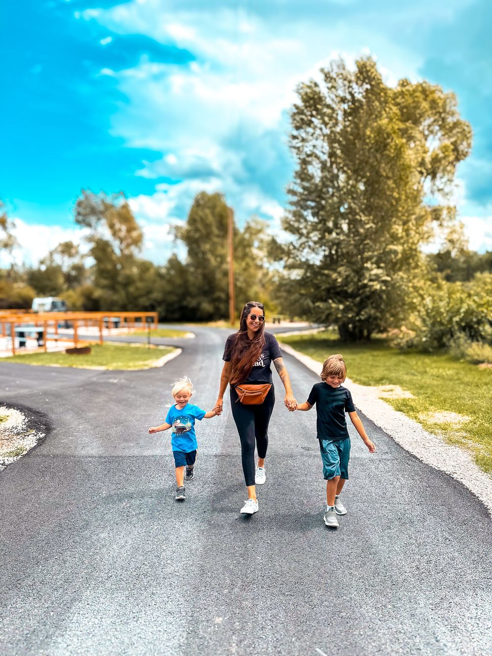 Woman and two children walking hand-in-hand down a sunny road lined with green trees and blue skies.