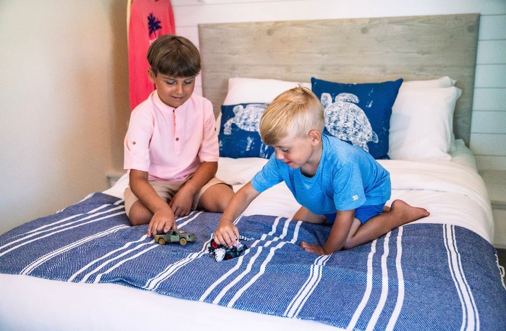 Two boys playing with toy cars on a striped blue bedspread in a cottage bedroom.