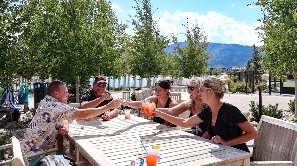 Group of four friends toasting drinks at outdoor table with mountain lake view.