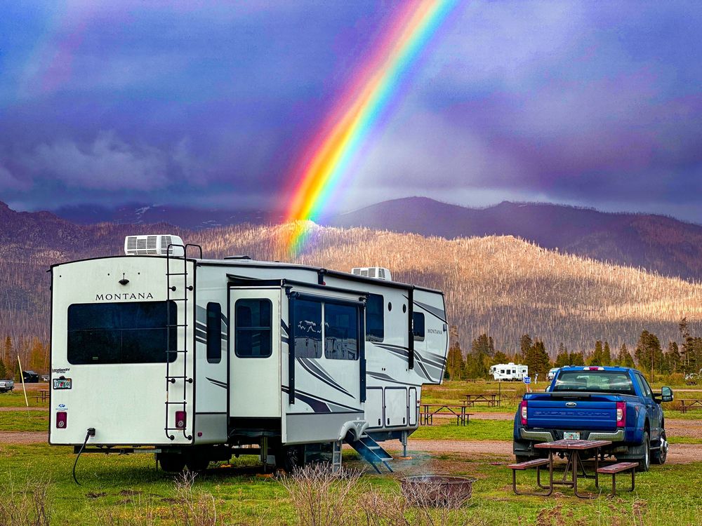 Fifth-wheel RV and blue truck parked at campsite with stunning rainbow over mountain backdrop.