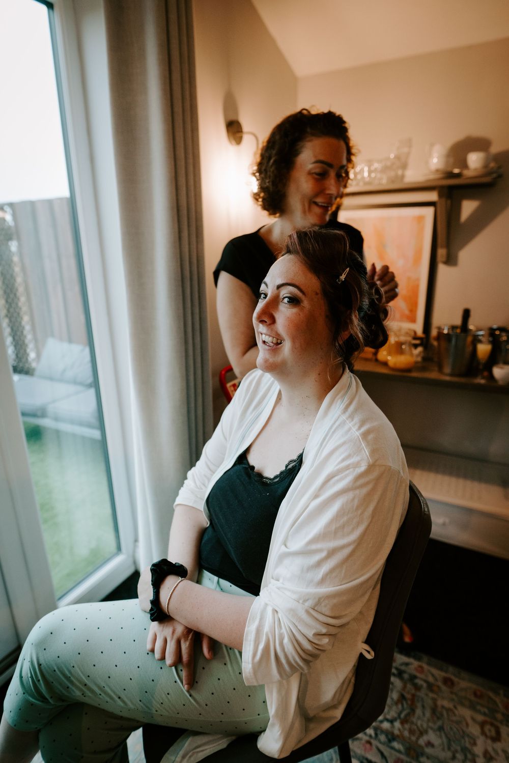 Bride smiling joyfully as hairstylist styles her hair by a bright window during wedding morning preparations.