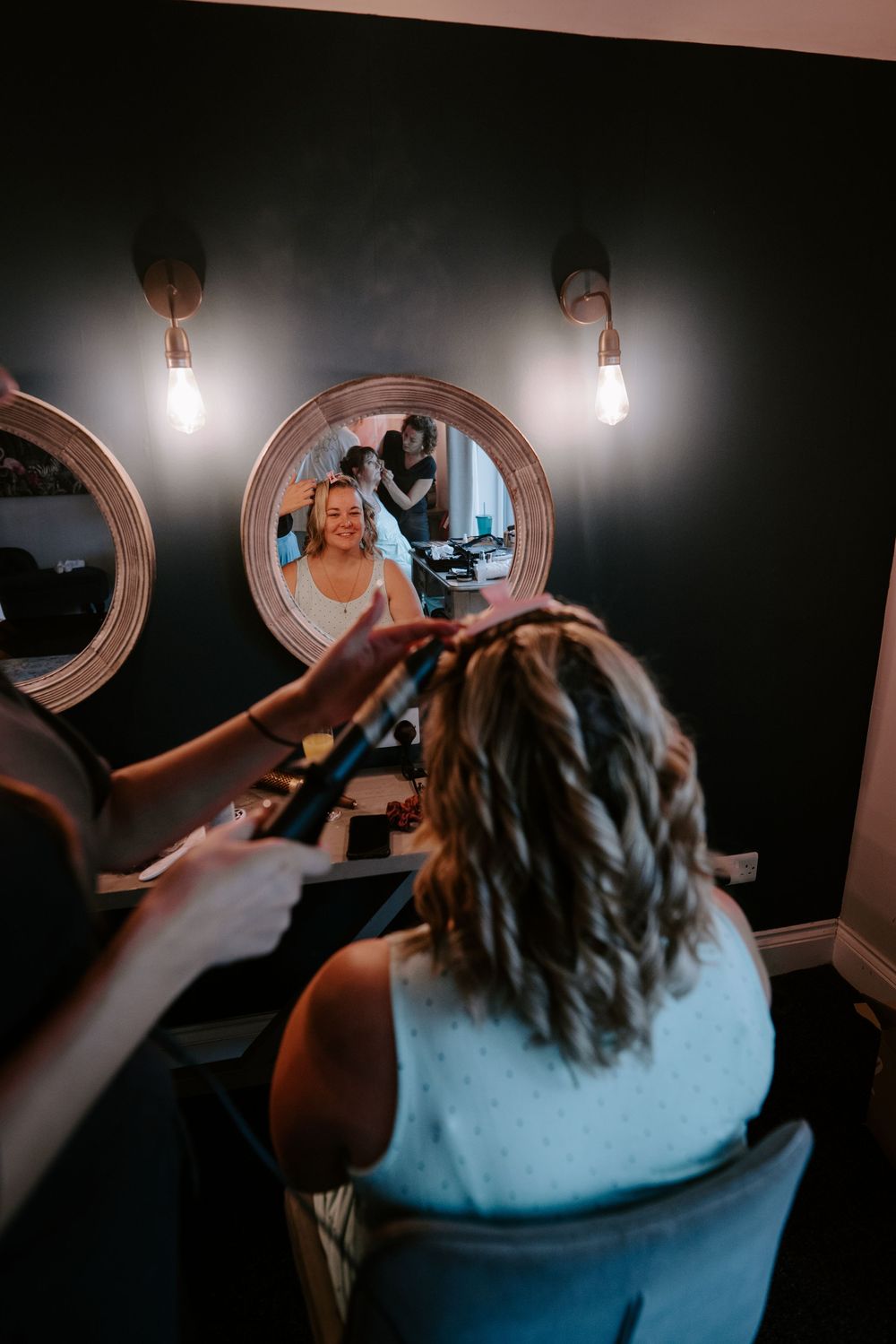 A hairstylist curling a smiling bride's hair at a vanity mirror in a dark-walled salon.