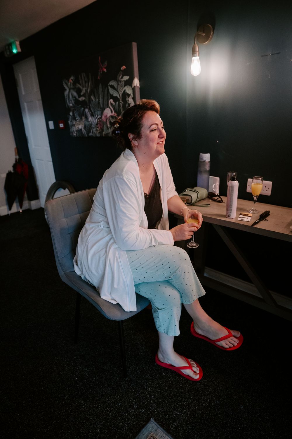 Bride relaxing in a white robe and flip flops, holding a champagne glass while getting ready for her wedding.